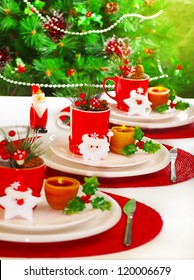 Picture of festive table setting, red cup for tea full of different Christmastime decoration, small Santa Clause decor with yellow candle on white white plate on Christmas tree background