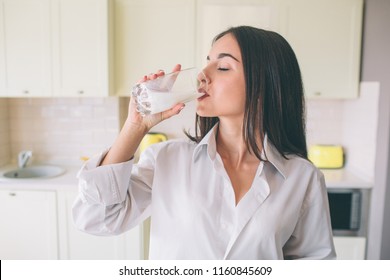 A picture of gorgeus girl standing in kitchen and drinking milk from glass cup. She is keeping eyes closed. Woman wears white shirt. - Shutterstock ID 1160845609