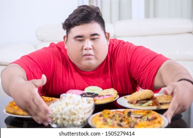 Picture Of A Greedy Fat Man Hugging At A Lot Of Unhealthy Food On The Table. Shot At Home