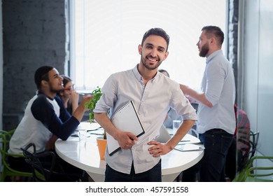 Picture Of Handsome Bearded Man In White Shirt Standing With His Hand On Hip. Executive Businessman Posing With Clipboard In Office Interior And Smiling.