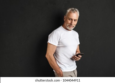 Picture Of Happy Handsome Mature Old Man Standing Isolated Over Black Wall. Looking Aside Using Mobile Phone.