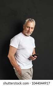 Picture Of Happy Handsome Mature Old Man Standing Isolated Over Black Wall. Looking Aside Using Mobile Phone.