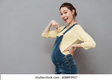 Picture of happy pregnant woman posing isolated over grey wall. Looking at camera and pointing.