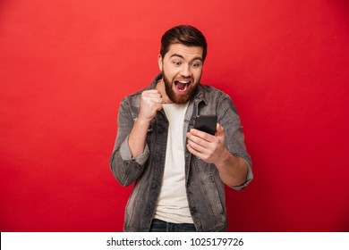 Picture of Happy screaming bearded man using smartphone and rejoices over red background