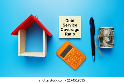 A Picture House Miniature, With Fake Money, Debt Service Coverage Ratio On Notepad, Calculator, And Pen On Blue Background.