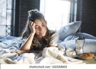 Picture Of Joyful Lazy Young Woman With Messy Hair Lying Awake In Bedroom After Good Sleep, Having Breakfast In Bed Made By Her Husband, Feeling Happy And Relaxed, Going To Drink Coffee With Croissant