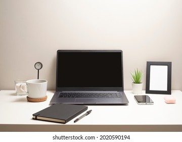 Picture Of Laptop On A White Table With A Mug A Clock A Small Bush A Notebook A Phone A Picture Frame And A Headphone Case