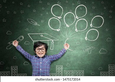 Picture Of Little Boy Looks Happy While Celebrating His Graduation And Holding A Diploma And Graduation Cap In Chalkboard Background