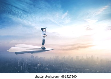 Picture Of Little Boy Standing On A Paper Plane While Flying Above City And Looking With Binoculars