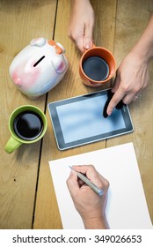 Picture Of Male Hand Writing On Paper And Female Hands Pointing On Tablet. Closeup Of Family Budget Or Project Planning With Cups Of Coffee On Wooden Plank Background.