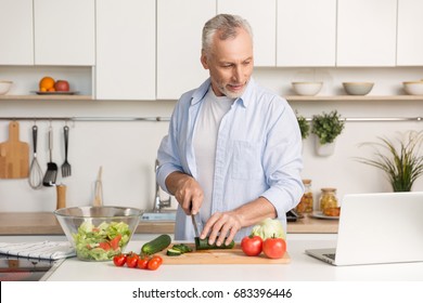 Picture of mature handsome man standing at the kitchen using laptop computer and cooking salad. Looking aside. - Powered by Shutterstock