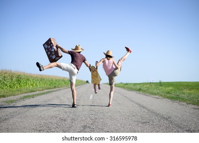 Picture Of Merry Family Jumping And Dancing On Road In Summertime. Backview Of Parents And Baby Girl With Retro Suitcase On Sunny Sky Countryside Background.