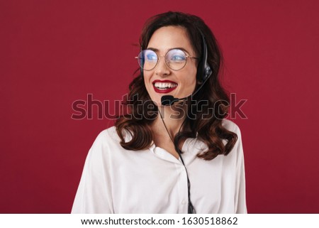 Picture of positive smiling young woman work in callcenter isolated over red wall background talking by cellphone.
