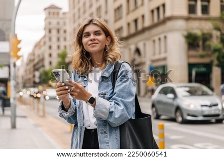 Picture of pretty young woman staying on the street holding phone in hands. Stylish blonde looking away and smiling in city. Use technology concept 