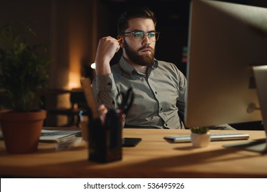 Picture Of Serious Bearded Web Designer Dressed In Shirt Working Late At Night And Looking At Computer While Touching Head With Pencil.