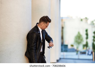 Picture of serious confident young man in stylish clothes leaning on white column outdoors and looking at his watch, checking time, getting annoyed while waiting for his girlfriend who is being late. - Powered by Shutterstock