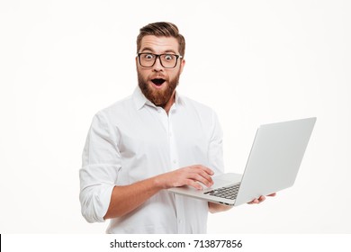 Picture of shocked young bearded man standing over white wall background isolated. Looking camera using laptop. - Shutterstock ID 713877856