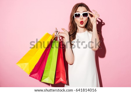 Picture of a shocked young brunette woman in white summer dress wearing sunglasses posing with shopping bags and looking at camera over pink background.