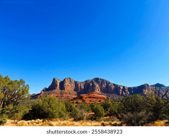 The picture showcases the majestic red rock formations of Sedona, Arizona, under a clear blue sky. In the foreground, a desert landscape stretches out, dotted with green shrubs and trees. - Powered by Shutterstock