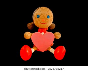 The picture shows a cute carved wooden doll of a girl with brown hair, blue eyes, orange nose, red mouth, two hands holding a pink heart, red shirt and pants, big red feet, beautiful colors. - Powered by Shutterstock