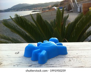 The picture shows a small blue sand toy a laughing crab lying on a wooden shelf. In the background you can see the leaves of a palm tree and a light sandy beach.
