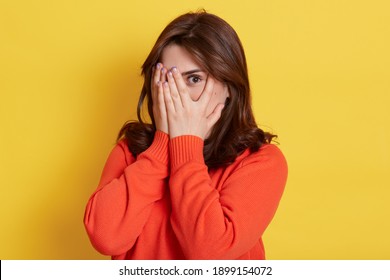 Picture of shy brunette teenage girl covering face with hands and peeping through her fingers at camera. Young attractive female hiding her face being frightened, scared or ashamed, wears casually.