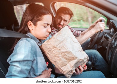 A picture of sick girl holding a bag and trying to vomit into it. She feels bad. Girl is holding her hand on stomach. Guy is looking at her with sight full of disgution.