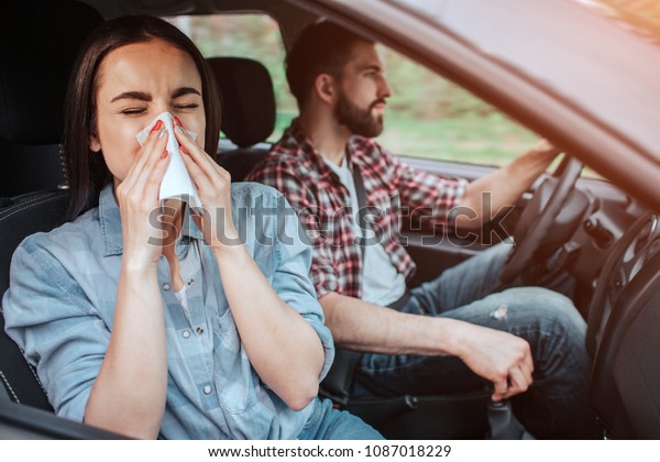 A picture of sick girl riding in car with\
young man. She is sneezing in napkin while he is paying attention\
to the road. Girl is\
suffering.