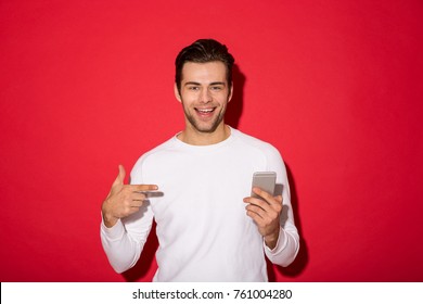 Picture of smiling man in sweater looking at the camera while holding smartphone and pointing at him over red background - Powered by Shutterstock