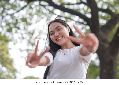 A Picture Of A Smiling Young Woman Holding Up A Peace Sign With Greenery In The Background.