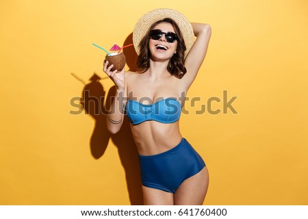 Picture of smiling young woman in swimwear isolated over yellow background holding cocktail.
