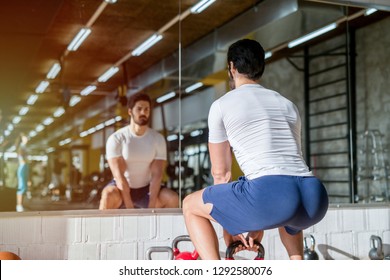 Picture Of Strong Focused Young Man Doing Squats With Weight In A Gym. Standing In Front Of The Mirror.