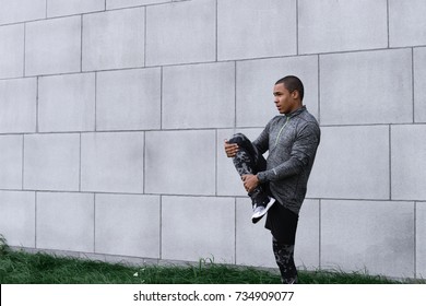 Picture Of Stylish Slim Young Dark-skinned Male Jogger Or Runner Wearing Leggings, Running Shoes And Fleece Jacket Warming Up Legs Outdoors Before Morning Cardio Workout Routine. Sports And Fitness