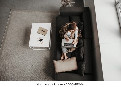 A Picture That Was Made From Above. Girl Sits On Balck Couch And Holds Laptop On Her Knees. She Has Blanket Around Her Body. Girl Works. There Are Phone And Notebook At White Table