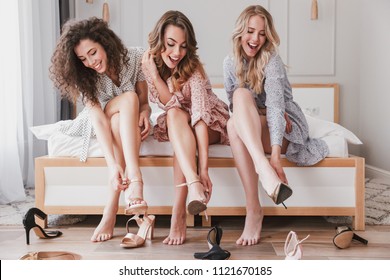 Picture of three gorgeous girls 20s wearing dresses trying on different summer stilettos or high heels during bachelorette party in posh bedroom