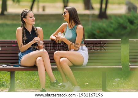 Picture through park bushes of two joyful young women drinking their smoothies while sitting on a bench outdoor