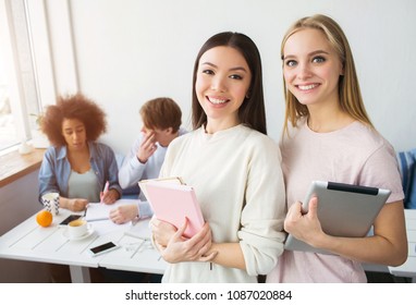 A Picture Of Two Beautiful Asian Girls Standing And Smiling. Brunette Holds A Pink Notebook While The Blonde Girl Has A Tablet In Her Hands. There Their Frinds Sitting Behind Them And Studying.