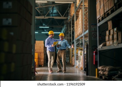 Picture Of Two Business Man Standing In Warehouse With Helmets On Their Heads And Celebrating Great News About Their Business. Standing In Big Warehouse And Looking Happy And Satisfied.