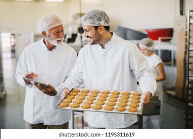 Picture of two employees in sterile clothes in food factory smiling and talking. Younger man is holding tray full of fresh cookies while the older is holding tablet and checking production line. - Powered by Shutterstock