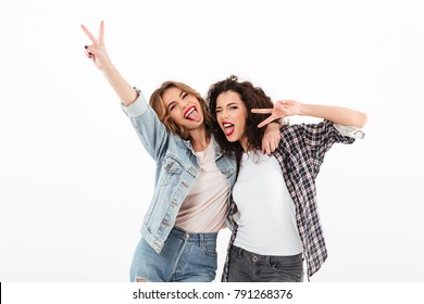 Picture Of Two Playful Girls Standing Together And Showing Peace Gestures While Looking At The Camera Over White Background