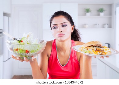 Picture of unhappy Indian woman looks confused while choosing a vegetable salad or hamburger in the kitchen
