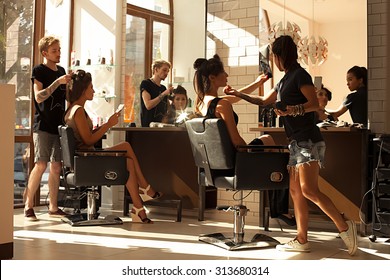Picture of working day inside the beauty- sit on two chairs clients beautiful young girls. Hairdresser makes hair styling or hair cut, make-up artist doing make-up in a beauty salon- stock photo.