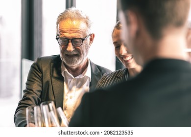 Picture Of Young Business Man And Woman Talking To His Older Business Partner. They Are In White Shirt And Black Tie. They Are Sitting On A Table In A Hotel Lobby. They Are Holding A Champagne Glass. 