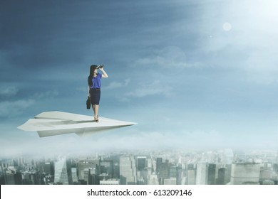 Picture of a young businesswoman looking through binoculars while flying with a paper plane above a city