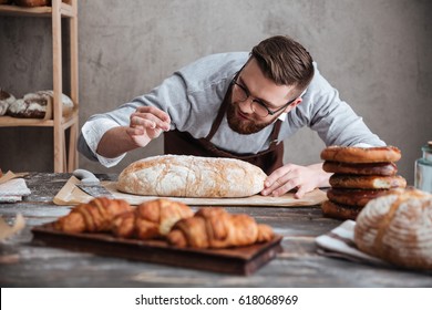 Picture of young concentrated man baker standing at bakery near bread. Looking aside.