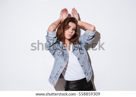 Picture of a young funny woman dressed in jeans jacket standing isolated over white background. Look at camera while showing bunny ears with hands.