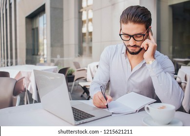 Picture of young man sitting at table and talking on phone. He is making notes in notebook and looks down. Businessman is serious and concentrated. There are laptop and cup of coffee at table. - Shutterstock ID 1215621211
