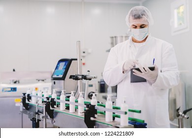 Picture Of Young Man In Sterile Clothes Using Tablet . Controlling Production Line On Factory. Bottles With Cosmetic Products On Production Line.