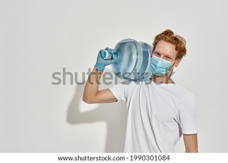 Picture of a young redhead deliveryman carrying a huge bottle of water on his shoulder wearing mask and gloves, front view, concept of safe delivery, stay at home