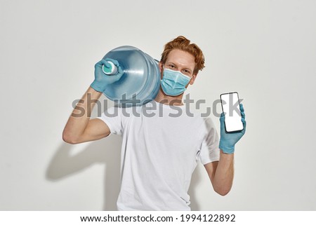 Picture of a young redhead deliveryman carrying a huge bottle of water on his shoulder, holding a smartphone wearing mask and gloves, front view, advertising app for water delivery